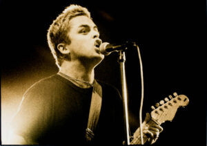 Lead singer and guitarist of the Band Green Day standing at a microphone singing and playing guitar in the 90's.