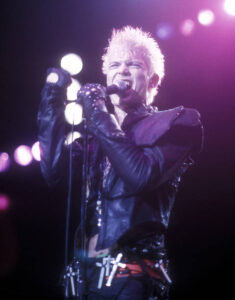 A young billy Idol performing on stage holding a microphone to his mouth singing.