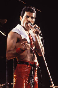 Freddie Mercury of Queen holding a microphone that is in a Mic stand to his mouth pointing his right hand while looking at the camera and singing