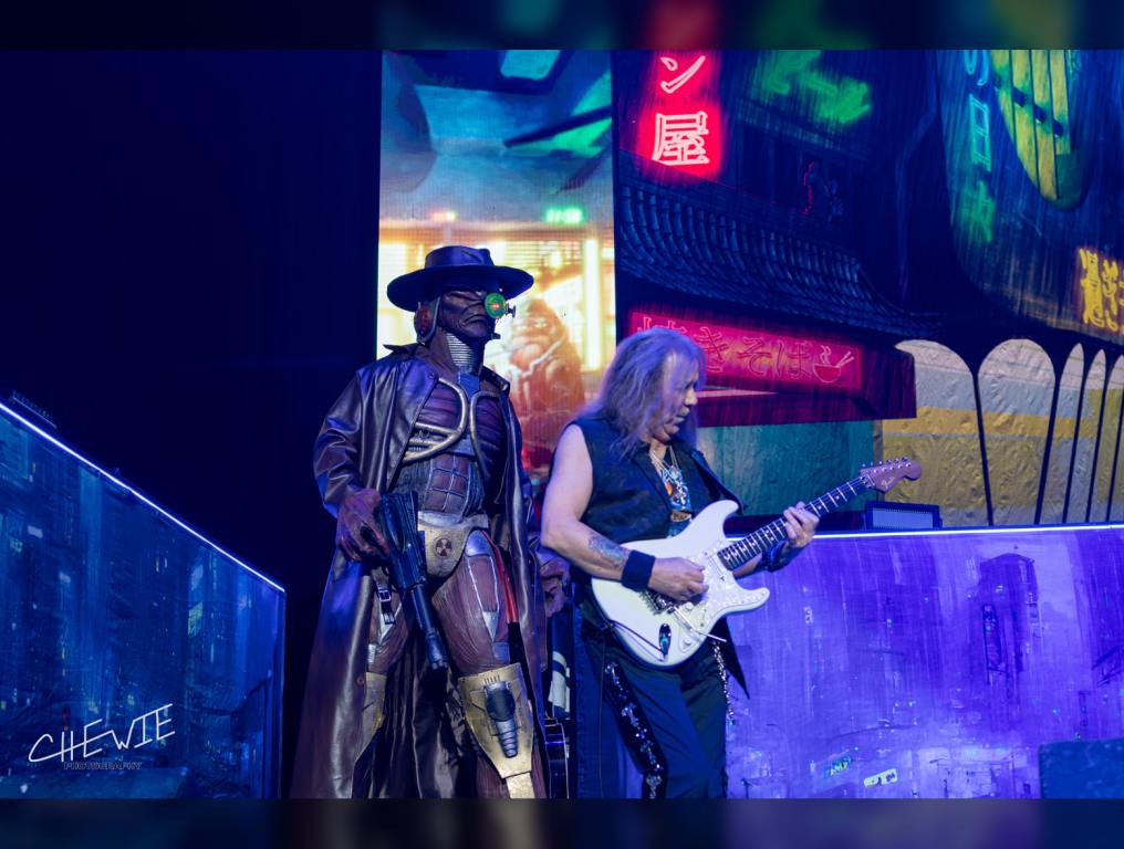 Iron Maiden mascot Eddie dressed in his "Stranger in a Strange Land" costume walks on the stage while guitarist Dave Murray plays in front of him.