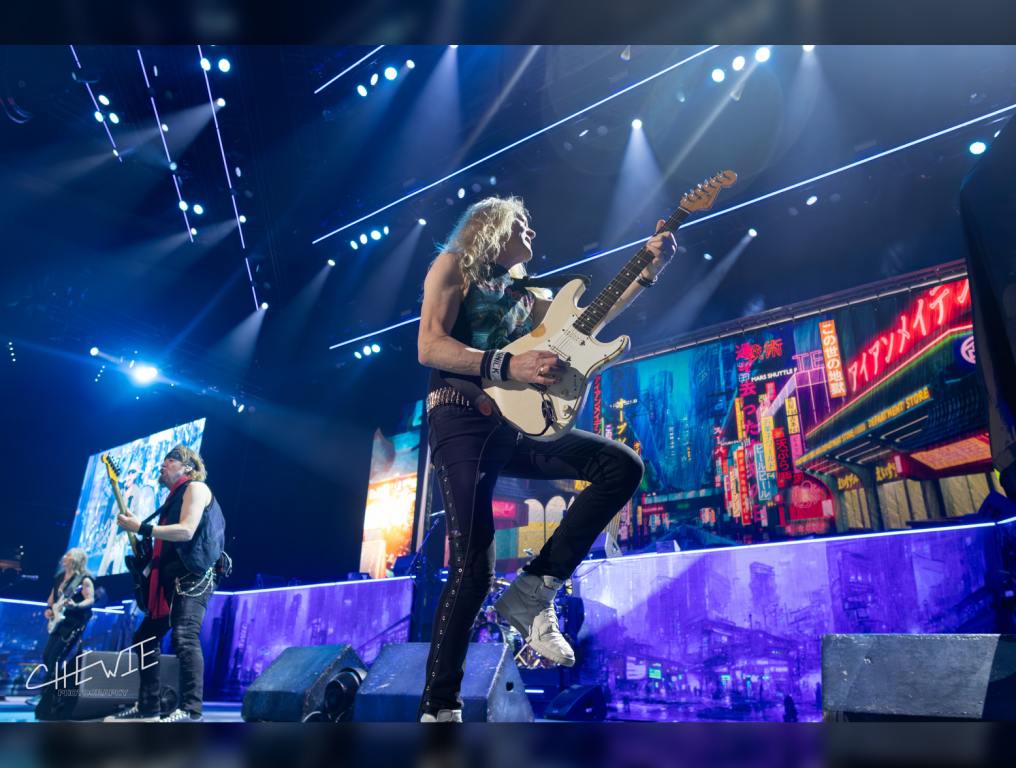 Iron Maiden's guitarist Janick Gers plays while holding his guitar up and fellow guitarists Dave Murray and Adrian Smith play in the background